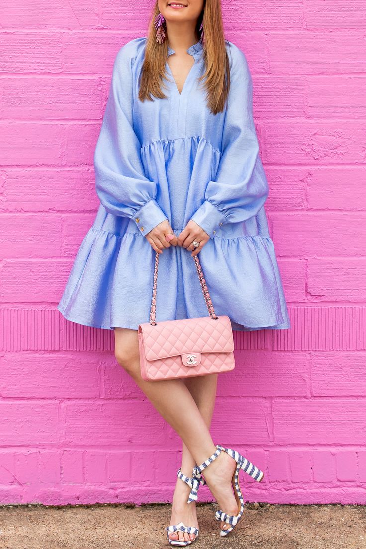 a woman is standing in front of a bright pink wall wearing a light blue smocked dress with puff sleeves. she is carrying a light pink chanel handbag and wearing navy and white striped highheeled sandals.  she has long brown hair and is wearing long tassel earrings. the overall style is feminine and casual.