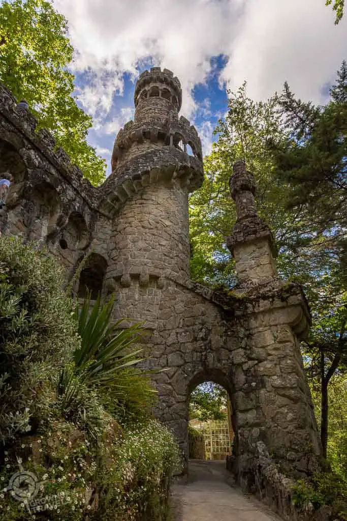 the image shows a stone tower and gate at quinta da regaleira in sintra, portugal. the tower is tall and cylindrical with decorative stonework. an archway forms the entrance to a pathway leading through a gate. lush greenery and trees surround the structure, and the sky is visible in the background with scattered clouds. the scene depicts a historical and architectural landmark.