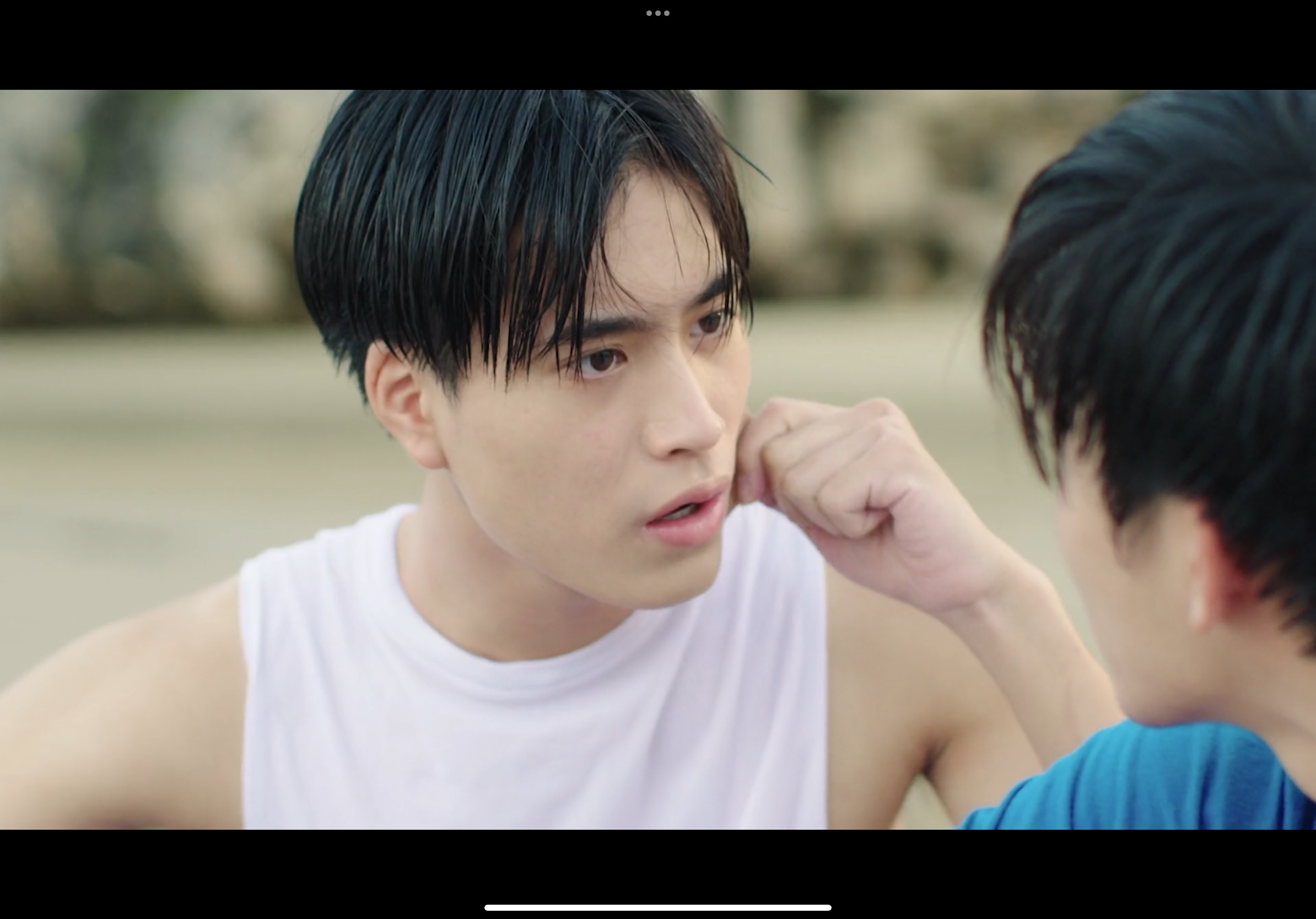 two young men are having a tense conversation on a beach. one man, wearing a white tank top, is looking intently at the other with an inquisitive expression. he has dark hair and a worried brow. the other man, partially visible and wearing a blue shirt, is turned towards the first man and has his fist raised to his chin, as if deep in thought. the background is a blurry, desaturated beach scene, suggesting an overcast day.  the overall atmosphere suggests a serious conversation, potentially about a conflict or emotional issue.