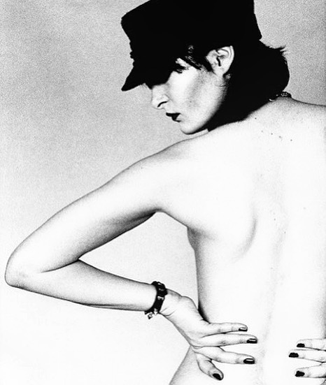 a black and white portrait of a young woman wearing a cap, with her hair covering most of her face. she is standing, and her arm is raised up with one hand on her back. the image is a studio shot, and the woman is looking away from the camera.