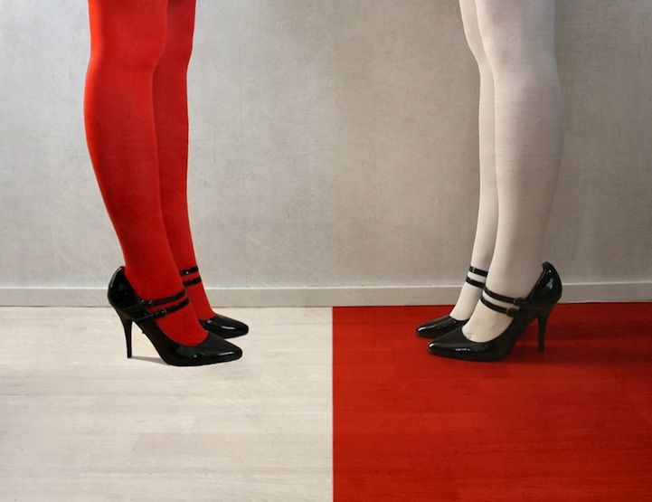 two women, one with white stockings and one with red stockings, stand with their legs apart wearing high heels.  the women are wearing identical black shoes with buckles. the floor is divided by a red and white stripe with the white stocking woman standing on the white side and the red stocking woman standing on the red side.