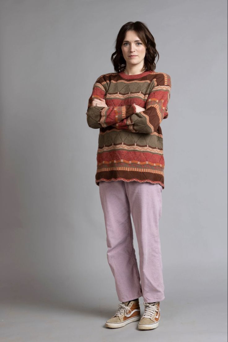 a woman stands with her arms crossed, wearing a patterned sweater, pink pants, and sneakers, against a gray background.