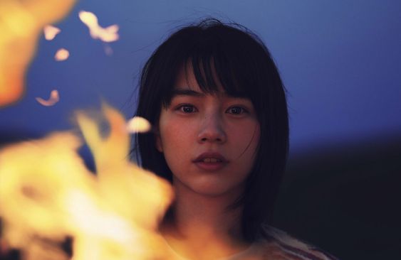 a young woman with dark hair and a serene expression is lit by a campfire in the foreground. the background is a blurry dusk sky and landscape.