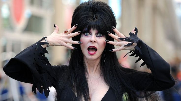 a woman with long black hair and dark clothing is smiling as she looks directly at the camera. she is wearing a long black dress and black gloves. her hair is styled in a beehive.