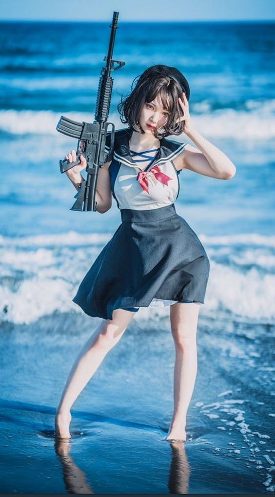 a young woman, likely cosplaying, stands on a beach with the ocean waves lapping at her feet. she is wearing a stylized sailor uniform with a short black skirt and white top accented with a red bow. she holds a black rifle in her left hand, resting it on her shoulder, while her right hand touches her hair. the overall tone of the image is blue and white, reflecting the colors of the sea and sky.