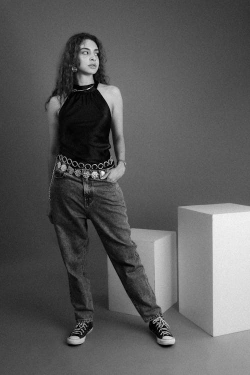 a black and white, fulllength studio shot of a young woman posing confidently. she's wearing a black halter top, loosefitting jeans, and converse sneakers.  her hair is styled in loose waves, and she has on several pieces of jewelry, including a statement belt. the backdrop is a simple grey studio with white cubic structures. the overall feel is stylish and modern.