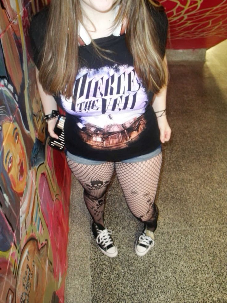 a woman is standing in a hallway, wearing a black "pierce the veil" tshirt, denim shorts, fishnet stockings with a skull pattern, and black converse sneakers. she is standing on a gray textured floor. to her left is a wall covered in colorful graffiti. the background also shows a glimpse of a red and white patterned wall.