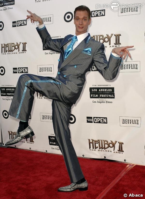 a man in a gray suit with a blue shirt and a blue tie is posing on a red carpet. he is smiling and has his left leg bent with his foot pointing to the left, and his right leg is straight. he is standing in front of a white wall with the words "hellboy ii: the golden army" in large black letters. there is a logo for netflix on the wall, along with several other logos.