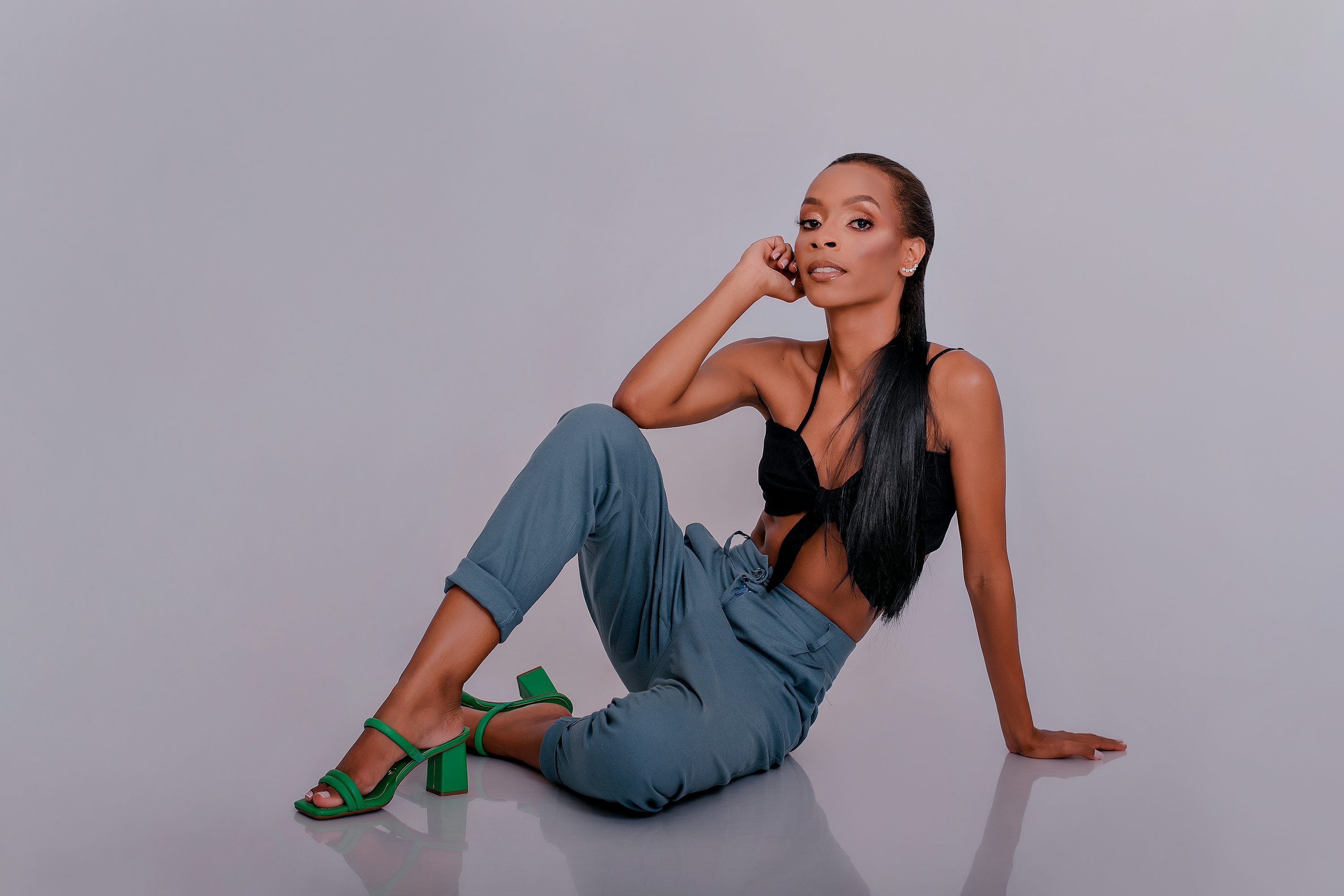 a young black woman with long dark hair is sitting on a grey background, posing for the camera. she's wearing blue pants, a black crop top and green highheeled sandals. her makeup is stylish, and her expression is confident and relaxed.  the overall image is fashionoriented and conveys a sense of style and elegance.