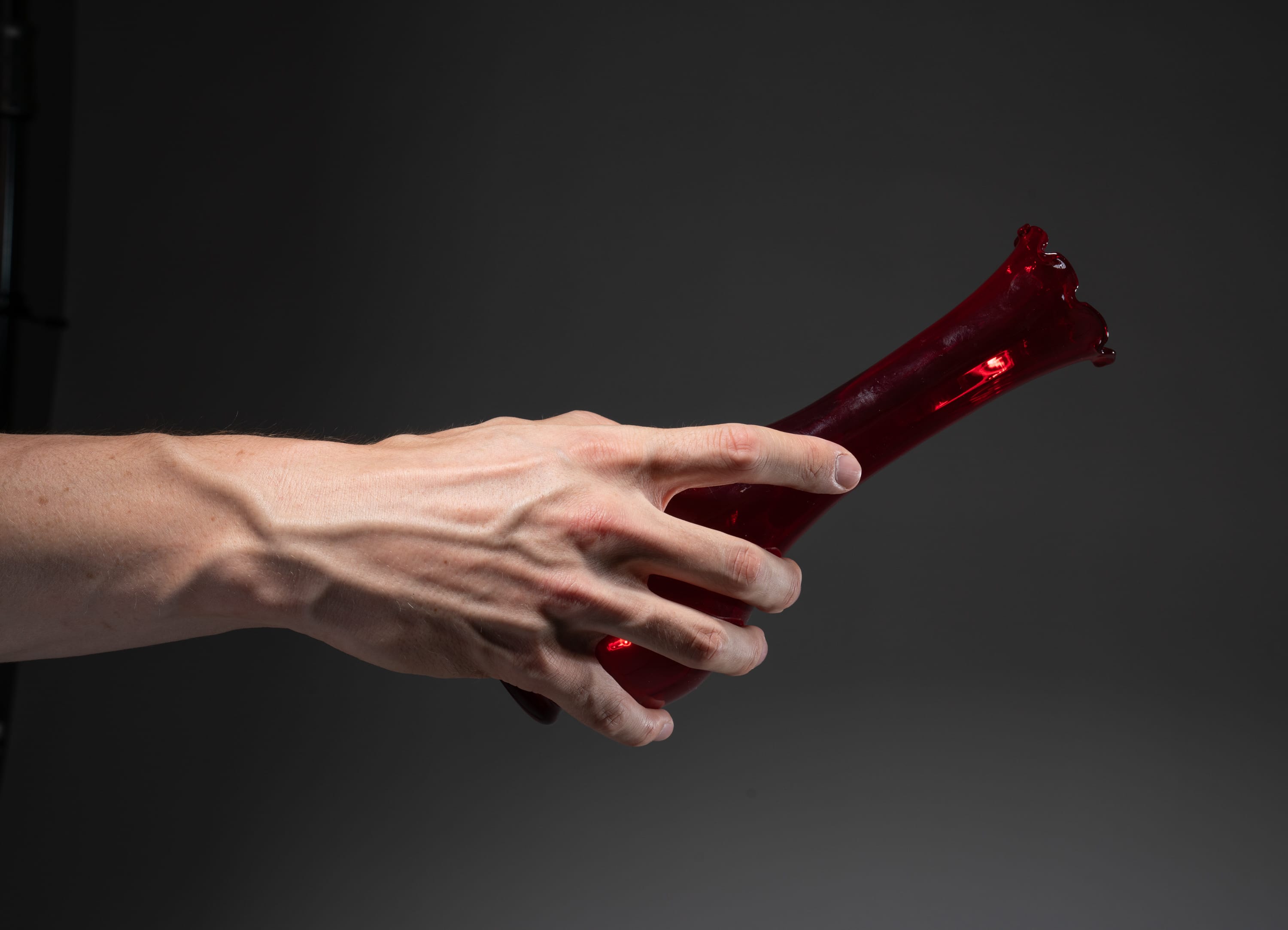 a caucasian left hand holding a red glass vase against a gray studio background. the vase is tilted as if the contents are about to spill out.