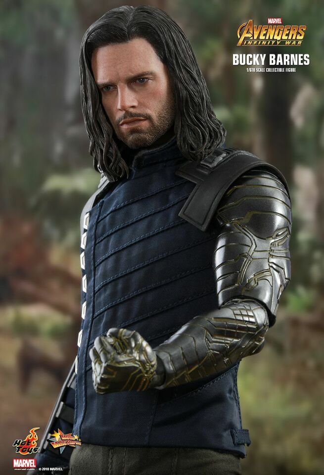 loseup image of a highly detailed 1/6 scale collectible action figure of bucky barnes from avengers: infinity war. the figure is wearing his signature dark blue vest with padded sections and his metallic prosthetic arm.  he has long dark hair and a serious expression on his face. the background is blurred, focusing the attention on the figure.  branding for hot toys and marvel is visible in the bottom corner.