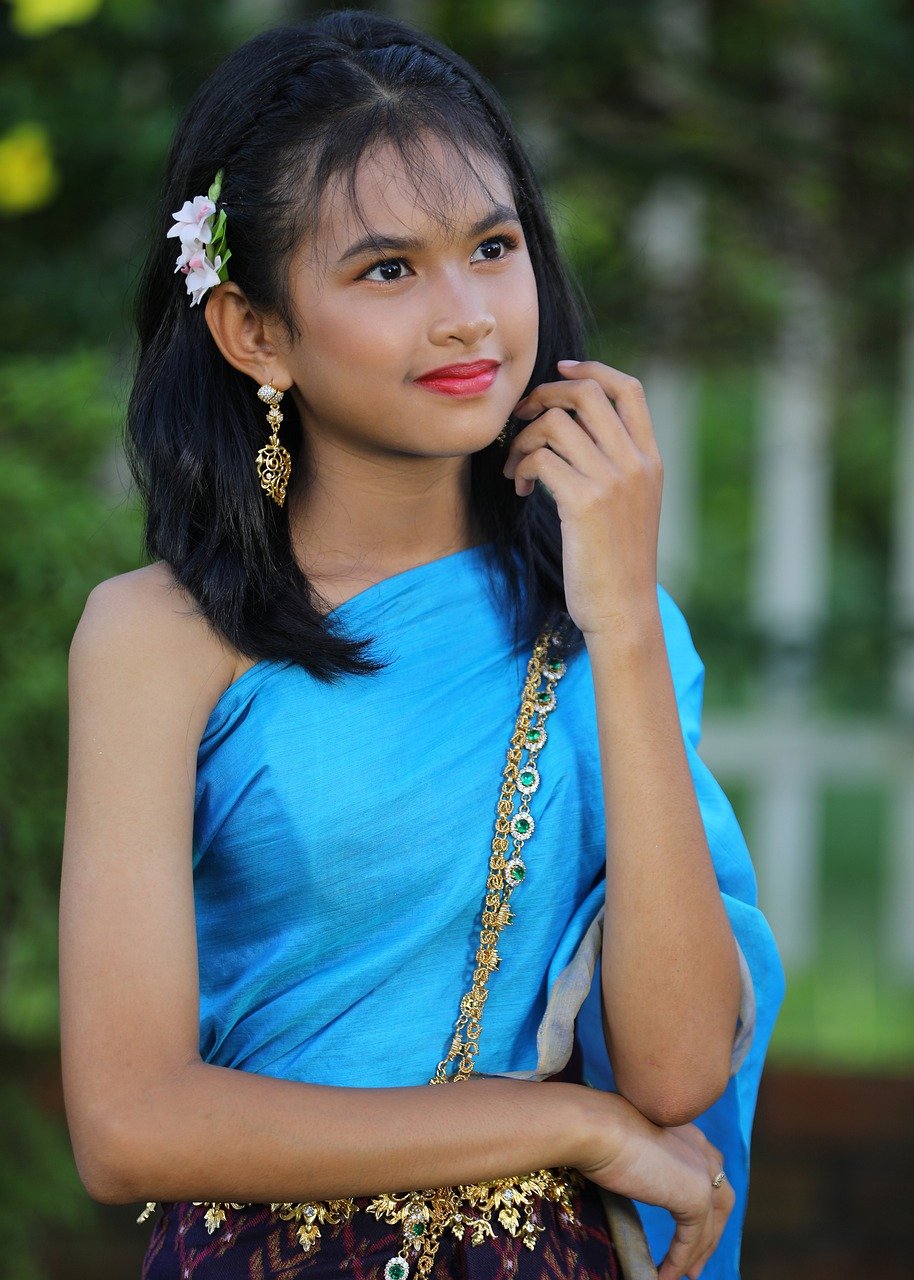 a closeup portrait of a young asian woman, possibly thai,  wearing a beautiful light blue traditional thai silk dress. she has a flower (possibly an orchid) in her hair and is adorned with gold and emerald green jewelry including earrings and a necklace. she's posing outdoors with a soft, natural light background. her expression is partly smiling and pensive, creating a serene and elegant mood. the image emphasizes her youthful beauty and the vibrancy of her traditional attire.