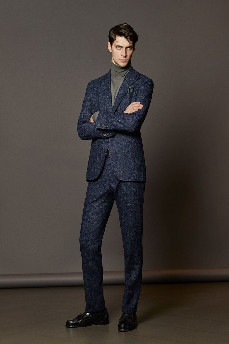 a fullshot studio portrait of a man wearing a blue tweed suit with a gray turtleneck sweater. he has his arms crossed and is wearing black leather shoes. he stands against a neutral gray background. a pocket square is visible in the breast pocket of the suit.