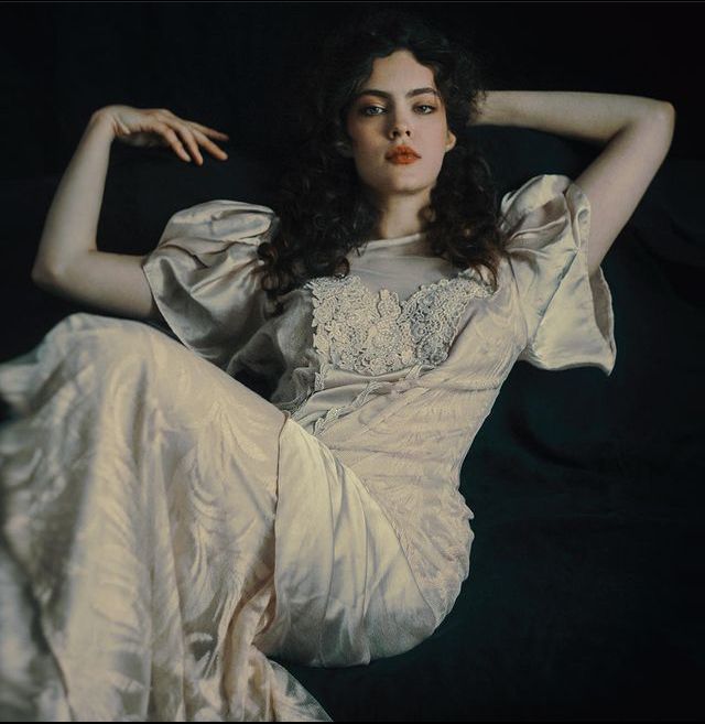 a woman with long, dark curly hair is sitting in a dark studio, looking directly at the camera with a hint of a smile. she is wearing a beautiful white lace dress with puff sleeves, which she looks stunning in. the background is dark and blurry to draw attention to the woman. the lighting is soft and romantic, and the overall effect is one of ethereal beauty.