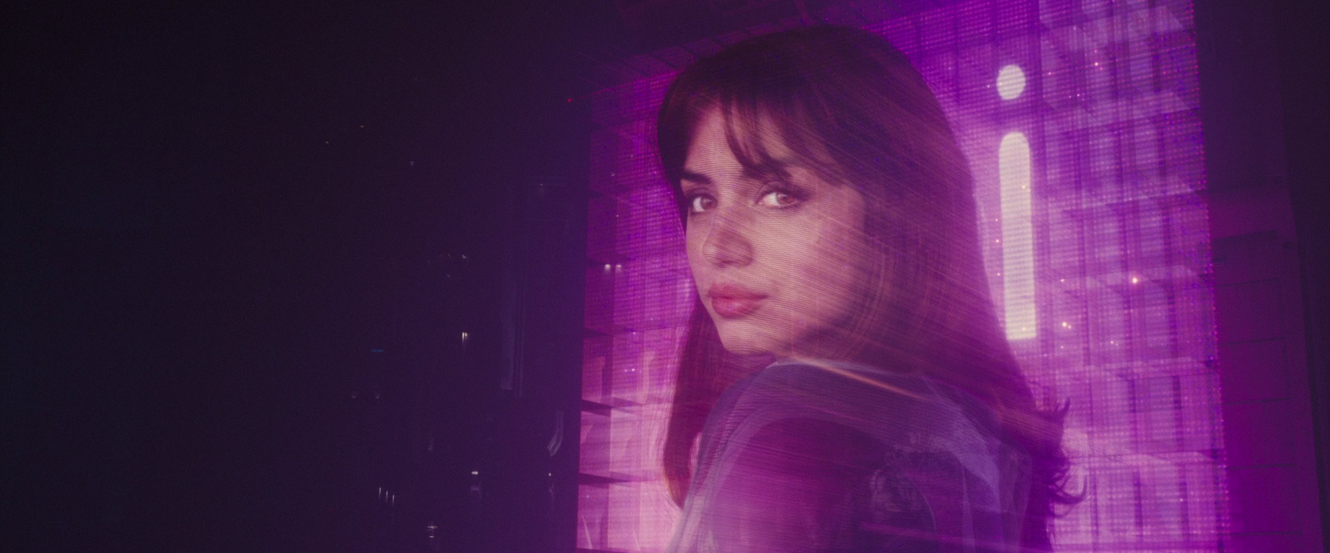 a woman with long brown hair and a serious expression stands in front of a wall of digital displays emitting a vibrant pink neon glow. the scene evokes a cyberpunk or futuristic aesthetic.