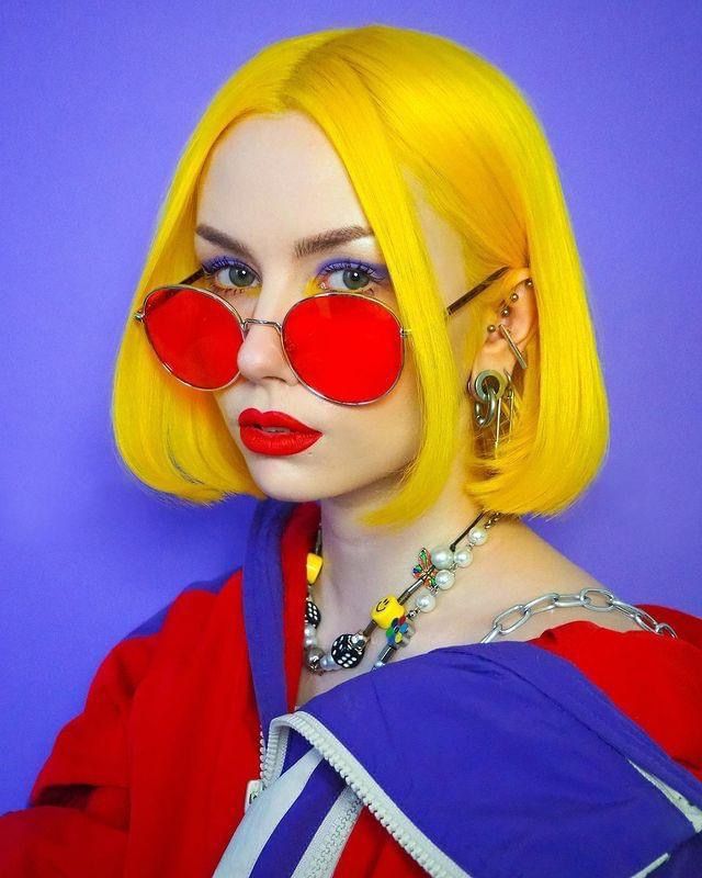 the image is a closeup portrait of a young woman with a bright yellow bob haircut. she's wearing large, round red sunglasses, vibrant red lipstick, and several necklaces including a pearl and charm necklace.  her jacket is a combination of red and purple. she also has visible piercings in her ears. the background is a solid, bright purple. the overall style is bold and retro, reminiscent of 90s fashion.  the image is sharply focused and welllit.