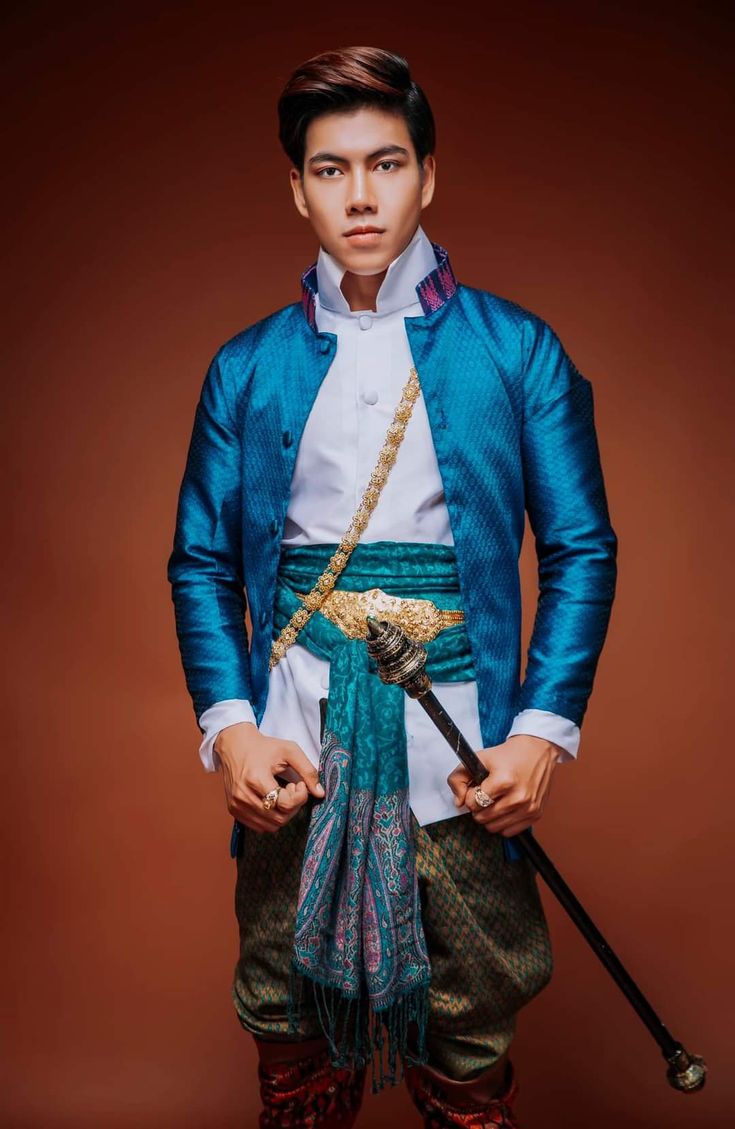 a handsome thai man in traditional clothing stands against a brown background and looks into the camera. he is wearing a blue jacket with a white shirt and gold chain underneath. he is wearing a blue and gold sash and a teal, gold and purple scarf around his waist. he is holding a traditional staff.
