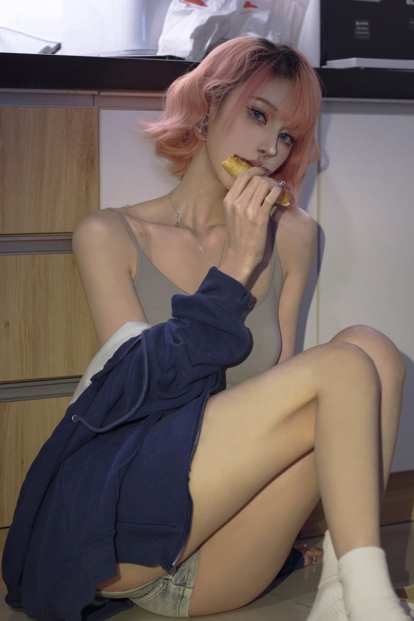 a young asian woman with short, pastel pink hair is sitting on a kitchen floor. she's wearing a light grey camisole, denim shorts, and a dark blue hoodie. she's eating a small piece of what appears to be pizza or a similar snack and is looking directly at the camera.  the overall style is soft and casual, with a focus on her delicate features and clothing. the image has a somewhat dreamy or softfocus quality.