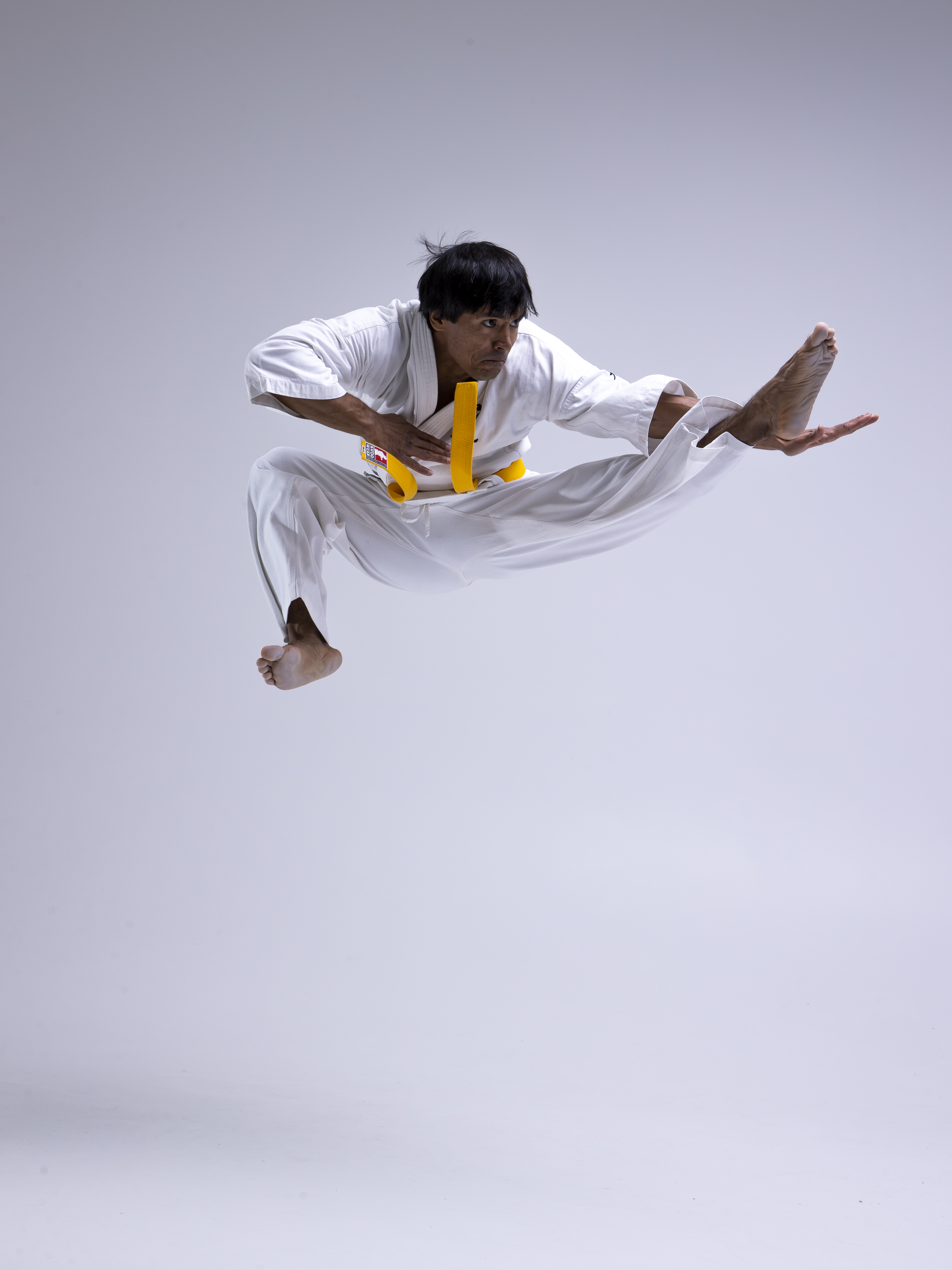 a man in a karate uniform with a yellow belt is performing a flying kick in midair. he is barefoot and wearing white sportswear. his legs are spread wide, and his arms are outstretched. the background is a plain white color. he seems to be performing karate or martial arts with motion.