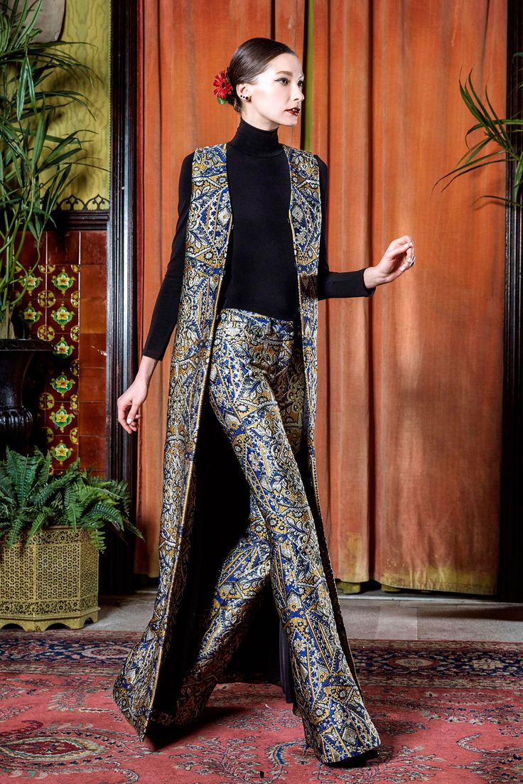 a fulllength shot of a fashion model showcasing a stylish outfit. she is wearing a long, patterned vest over a black turtleneck and highwaisted flared pants, all in coordinating rich blue and gold paisley brocade. the outfit is elegant and reminiscent of vintage 60s or 70s styles. the model is posing gracefully in an indoor setting with ornate curtains and a patterned carpet in the background.  she has her hair in a chic updo and minimal jewelry.  the overall image is a sophisticated fashion editorial.