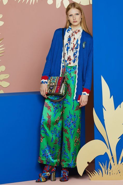 a fulllength shot of a fashion model posing against a colorful backdrop. she is wearing a bright blue cardigan over a white floral blouse, vibrant green floral wideleg pants, and patterned highheeled shoes. she carries a patterned crossbody bag. the background features shades of blue, pink, and beige, with cutout leaf shapes. the overall style is vibrant and summery.