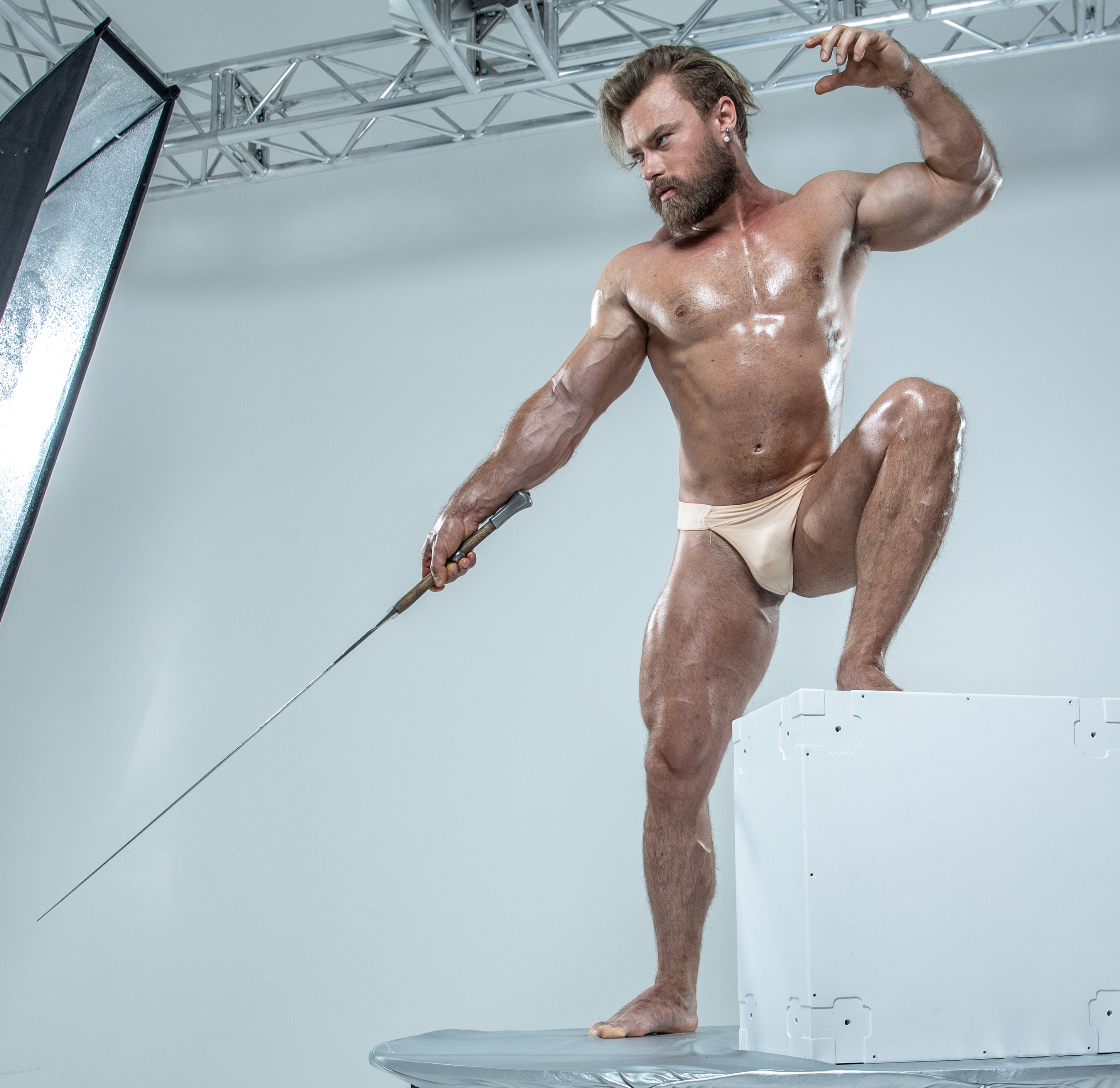 the image showcases a muscular man in a dynamic pose. he is holding a sword and standing on a white cube with his left foot. the background and the use of artificial lighting suggest a studio environment. the low camera angle emphasizes his powerful physique.