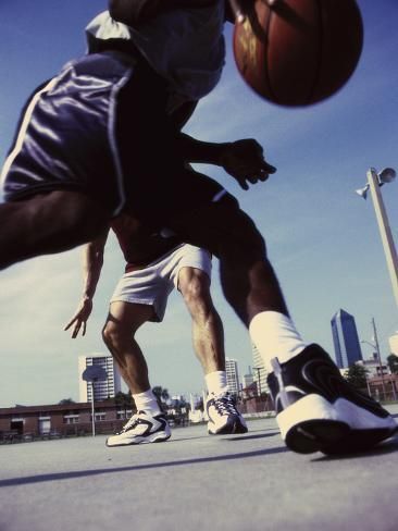 the image is a dynamic, lowangle closeup shot of two basketball players in action on an outdoor court. the foreground is dominated by the legs and feet of the player in the foreground, who is dribbling the ball. the other player is partially visible, appearing slightly blurred due to motion.  both players are wearing basketball shorts and athletic shoes. the background reveals an urban setting with buildings and a clear sky. the overall impression is one of speed, competition, and the energy of a basketball game. the image is sharply focused on the players' lower bodies and the basketball, with the background slightly out of focus.