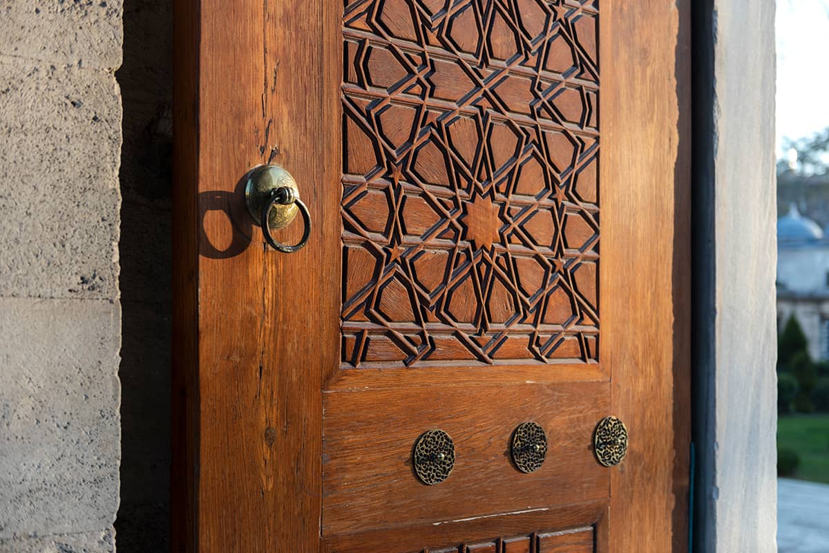 a closeup shot of a wooden door with intricate carvings and metal hardware. the door has a geometric pattern in the upper part and three circular metal ornaments in the lower part. a metal ring hangs from a knob on the left side of the door. the door is set into a stone wall.