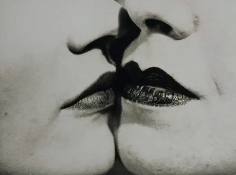 the image is a closeup black and white photograph of two women's faces, their lips almost touching in a kiss.  the focus is intensely on their mouths; dark lipstick is prominent.  the overall mood is intimate, sensual, and somewhat dramatic due to the contrast and dark tones. the style is reminiscent of classic vintage photography.