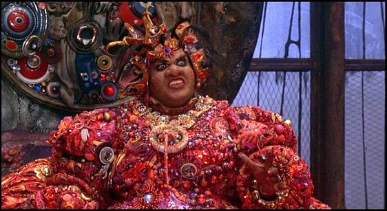 a woman wearing an ornate costume, jewelry, and a crown in a movie. the woman is in a dramatic pose and she appears to be an evil queen.
