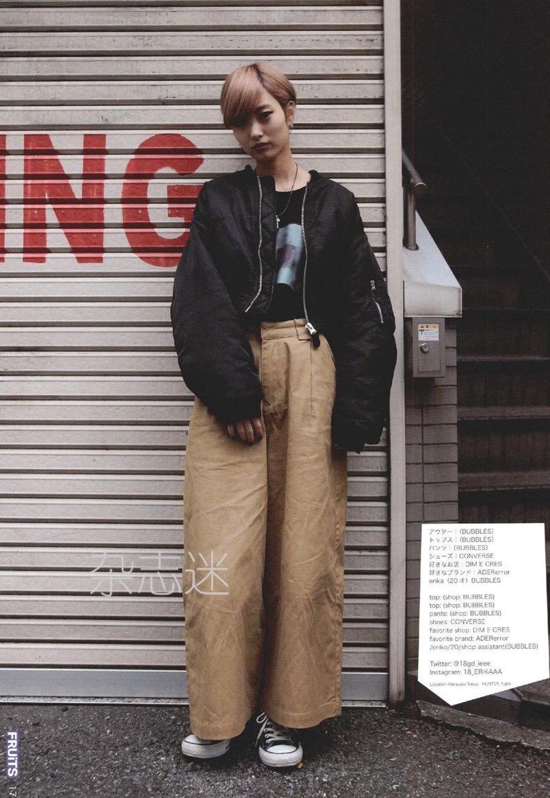 a young woman poses for a street fashion photograph. she is wearing a black bomber jacket, black tshirt, wide leg brown pants, and black and white converse. the background is the metal door of a storefront, and there is japanese text to the right of the image.
