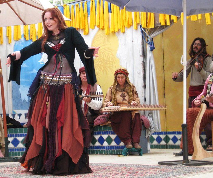 a woman with red hair performs belly dancing while a band plays stringed instruments behind her, all set on a stage decorated with a yellow streamer backdrop and blue and white trim, suggesting a festival setting.