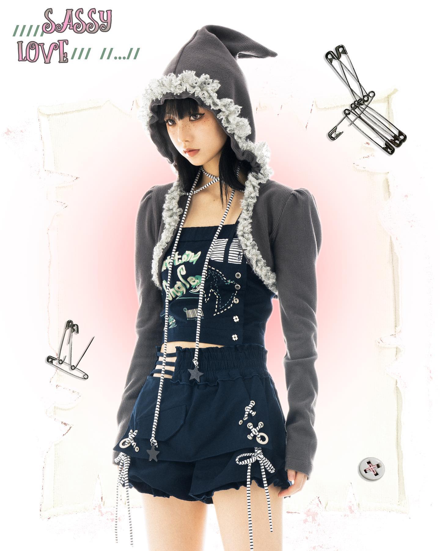 a young woman poses in a stylish outfit, which includes a dark gray hoodie with lace trim, a matching cropped cardigan, a dark blue layered shorts with star and lace accents, and a graphic printed top.  the image has a slightly gothic and kawaii aesthetic, accented by safety pins and a light pink background with design elements. the overall style suggests a blend of japanese street style and lolita fashion. the model's pose and expression are confident and fashionable. the word "sassy love" is written in stylized text at the top.