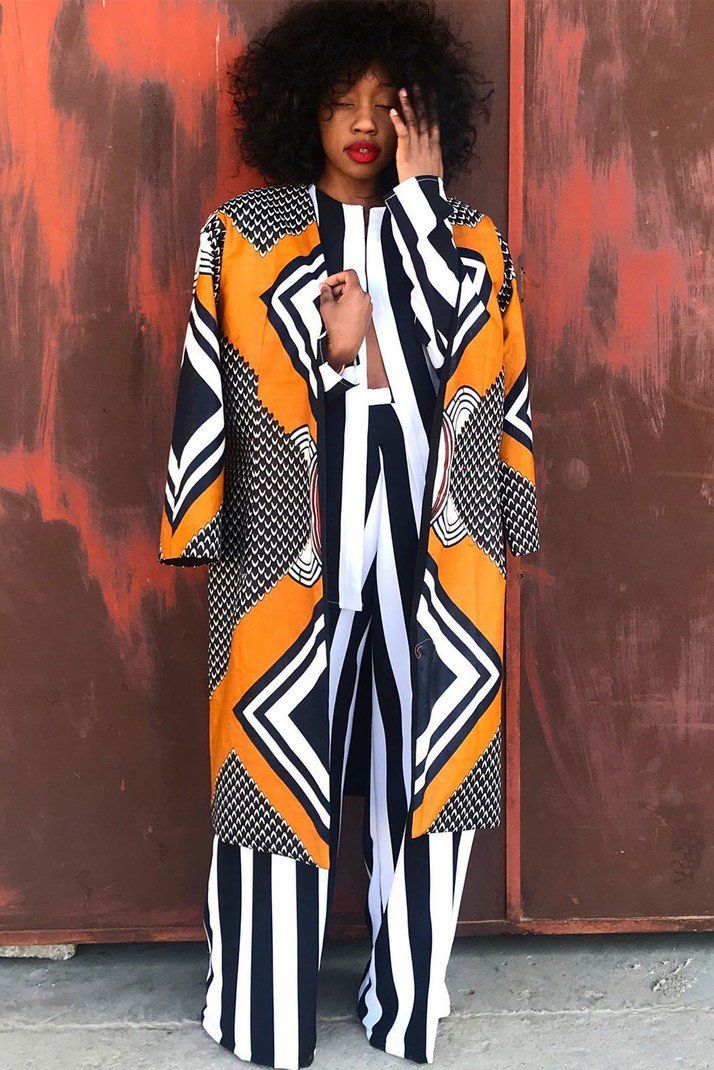 a woman with curly hair is standing in front of a rusty orange metal door. she is wearing a stylish longsleeved coat and palazzo pants, both in a vibrant african print featuring a mix of orange, black, and white geometric patterns. the coat is open, revealing a white and black striped shirt underneath. her pose suggests a contemplative or introspective mood. the overall image is a striking blend of bold colors and patterns against a textured backdrop.