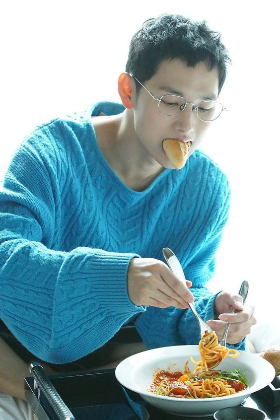 the image shows a man wearing glasses and a blue sweater sitting down and eating a plate of spaghetti. he has a piece of bread in his mouth and is twirling the pasta with a fork.