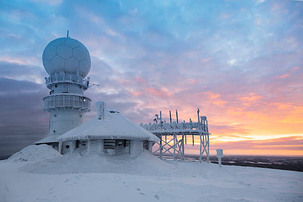 a snowcovered weather station stands on top of a mountain against a stunning sunrise. the sky is a mix of pink, orange, and blue hues, with fluffy clouds scattered across the canvas. the snowladen landscape stretches as far as the eye can see. the image captures the beauty and tranquility of a winter wonderland.
