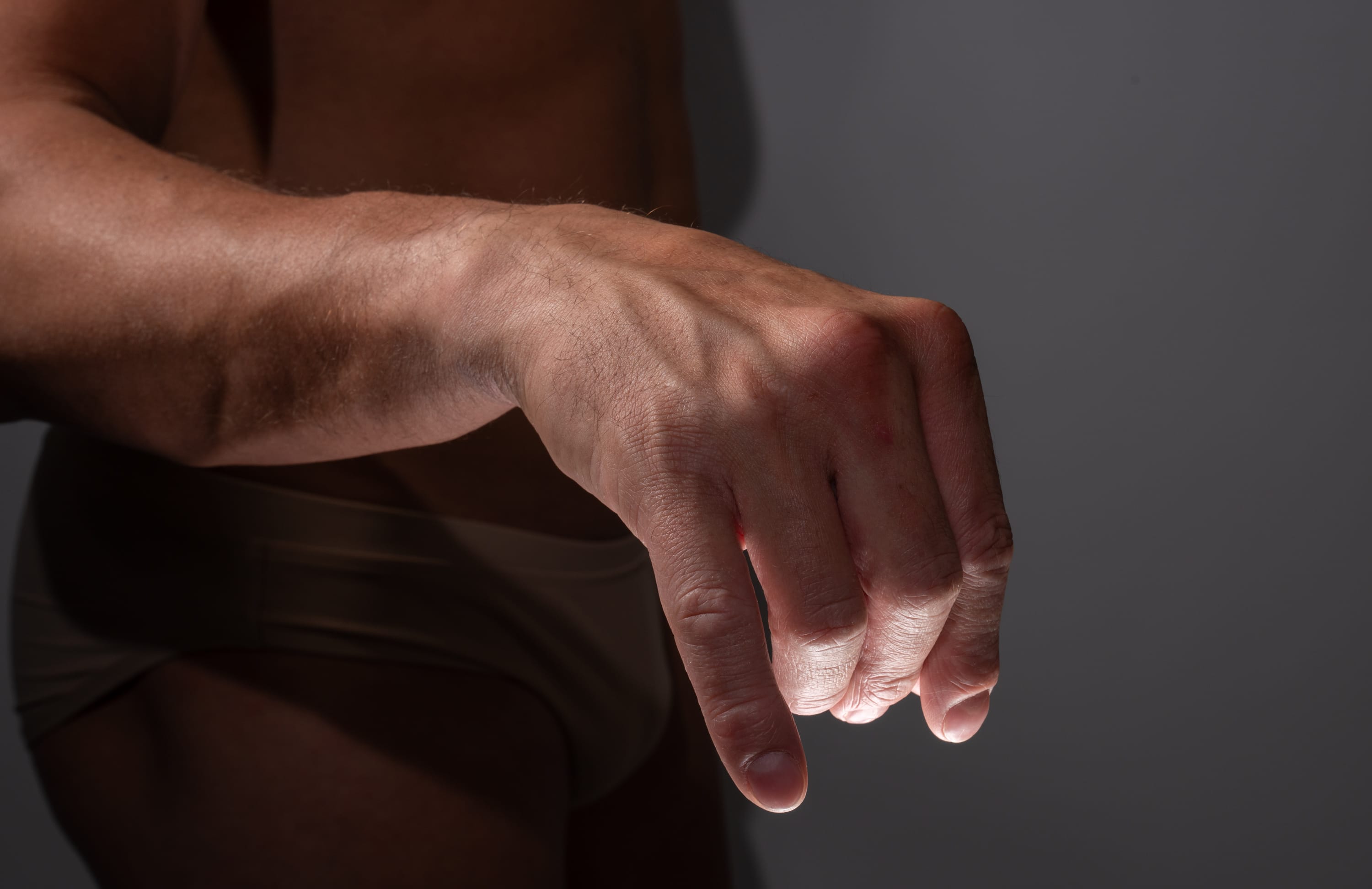 closeup shot of a muscular man's arm and hand extended forward against a dark gray background. the hand is partially clenched, with the fingers curled inward.  the lighting is dramatic, highlighting the contours of the arm and hand. the man is wearing tan underwear, which is partially visible in the frame.