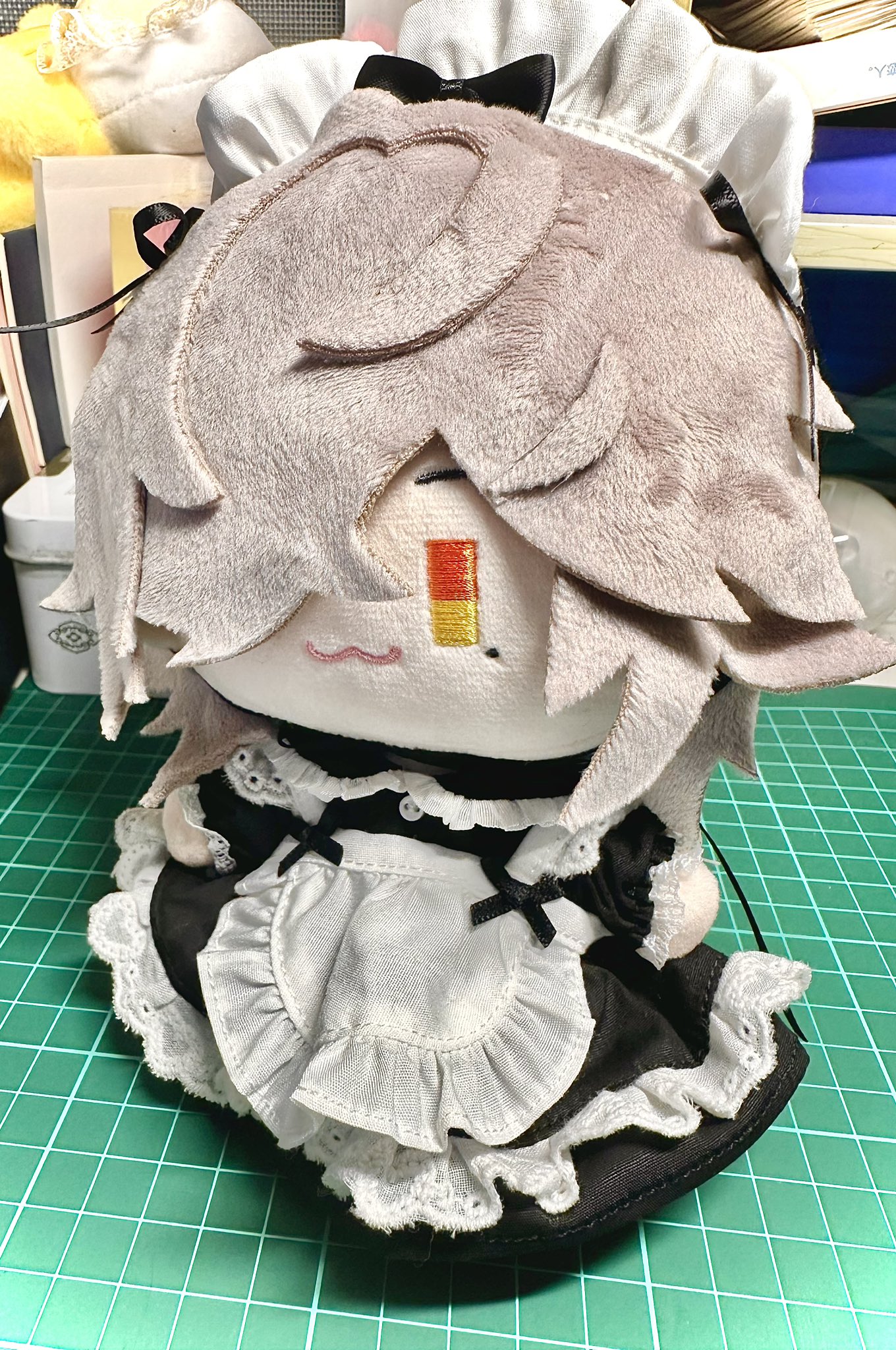 a closeup shot of a cute plush toy dressed in a maid outfit. the toy has gray hair and gray eyes and is wearing a white dress with a black lace trim and a black bow. the toy is also wearing a white apron with a black lace trim and a white bow. the toy is sitting on a green mat.