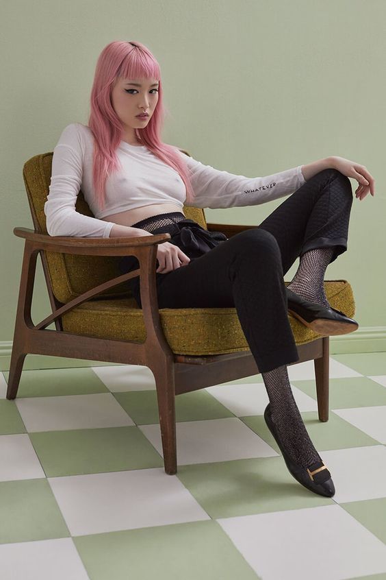 a woman with pink hair is sitting in a chair. she is wearing a white shirt, black pants, black shoes, and fishnet tights. the floor is green and white checkered. she has a tattoo on her arm that says "whatever".