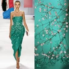 a fashion model stands on a runway wearing a sparkling green dress with a floral print. she is wearing her hair down, and she is holding a white clutch purse. the background is a blur of light blue and white, and there is a floral pattern on the right side of the image.
