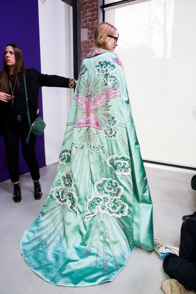 a model wears a long, embroidered cape backstage at a fashion show. the cape is light blue with a pink bird design on the back, surrounded by clouds. a woman with dark hair stands to the left, wearing a black top, black pants, and high heels. she also has a green bag slung over her shoulder. the model wearing the cape has short blonde hair and is wearing sunglasses.