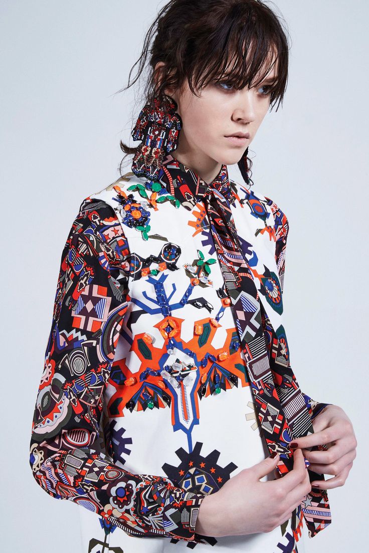 closeup shot of a female fashion model showcasing a vibrant, eclectic outfit.  the attire consists of a longsleeved shirt and a sleeveless top, both featuring a busy, multicolored pattern incorporating geometric and ethnic design elements. the colors are bold and include oranges, blues, reds, and blacks on a primarily white background.  the model is wearing large, ornate statement earrings that complement the outfit's bold style. her dark brown hair is styled with bangs, and she has a serious yet fashionable expression. the background is a plain light gray, focusing attention on the model and her clothing. the overall impression is one of high fashion and a contemporary, artistic approach to design.
