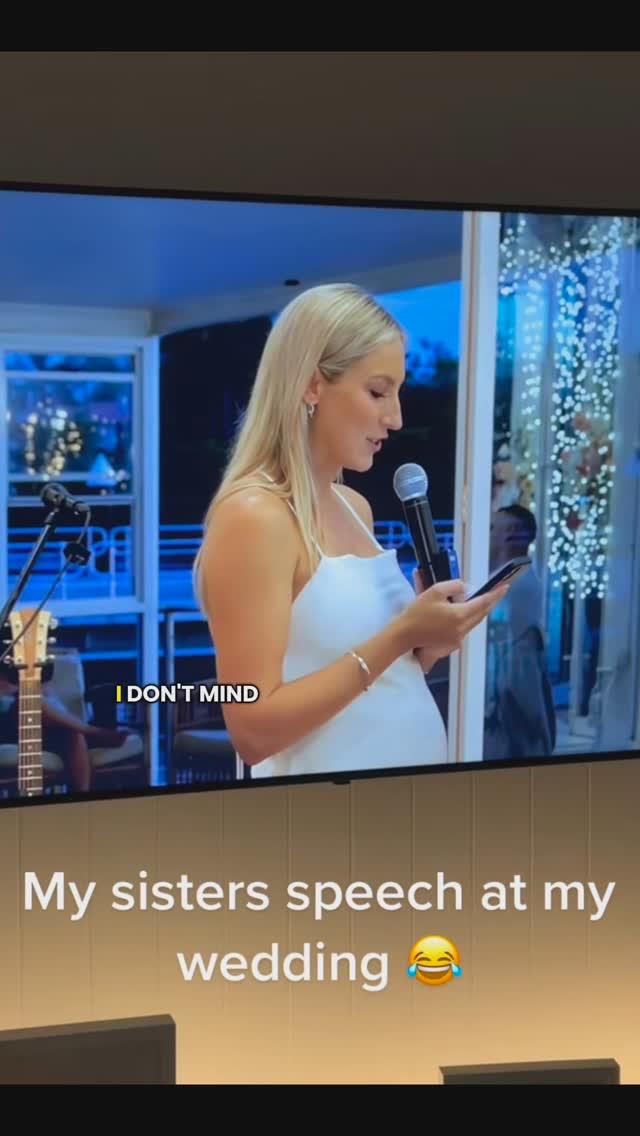 a photo of a screen displaying a wedding scene where a woman is giving a speech. the woman is blonde, wearing a white dress, and holding a microphone and a phone. she is standing on a stage with instruments visible in the background. text on the screen reads "i don't mind" and "my sisters speech at my wedding" with a laughing emoji.