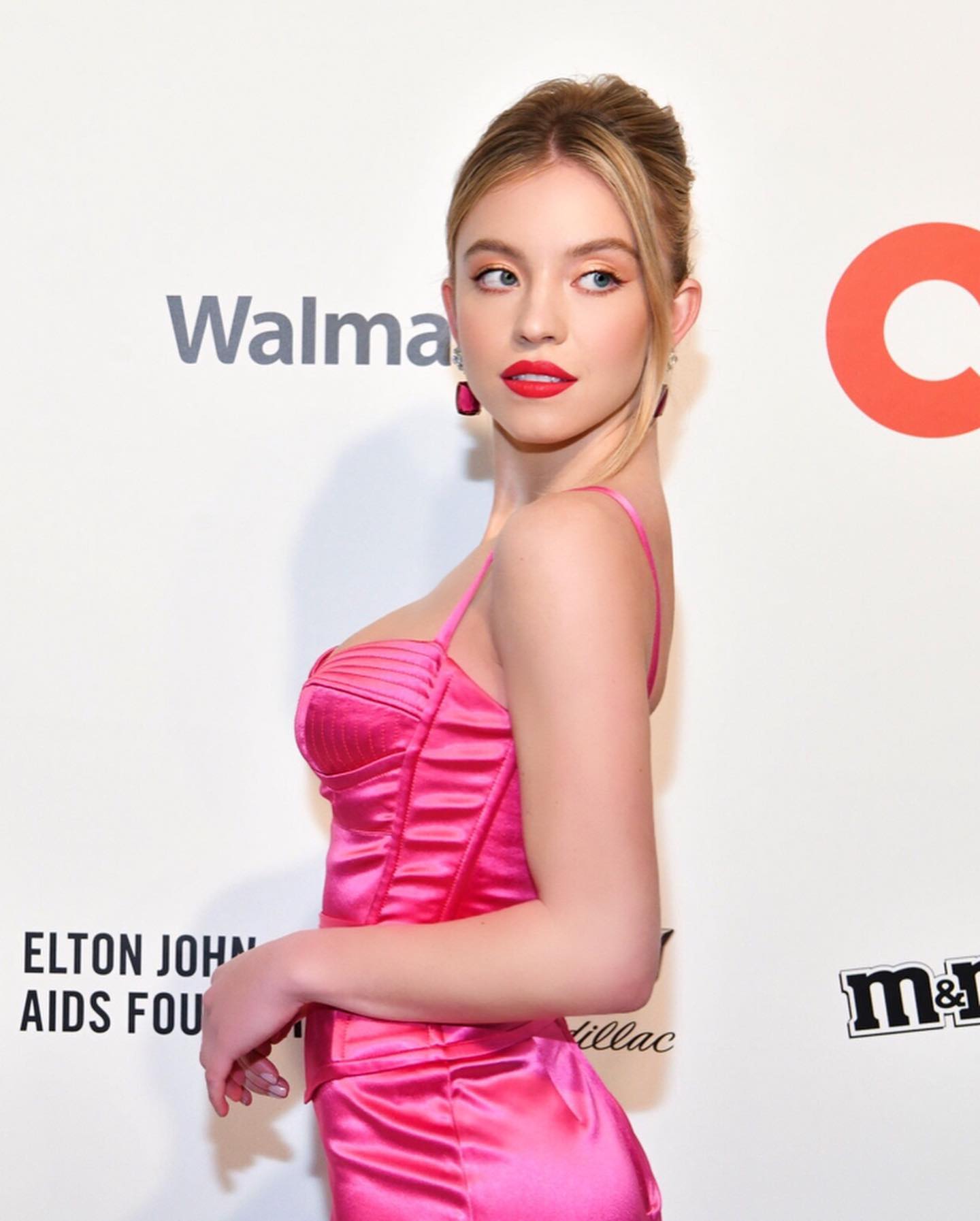 the image shows a woman, possibly an actress or model, at what appears to be an awards ceremony or gala event. she is wearing a vibrant hot pink satin corsetstyle top and matching skirt. her hair is styled in a sleek high bun, and she has on red lipstick.  the background features logos for sponsors, including walmart, cadillac, m&m's, and a mention of the elton john aids foundation. the overall feel of the image is glamorous and stylish. the photo is a profile view of the woman, showcasing her pose and the detail of her dress.