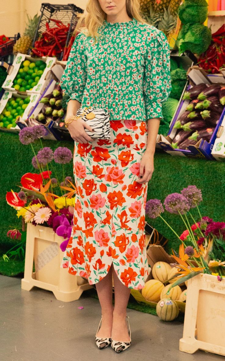 a woman is standing in a market setting, showcasing a fashionable outfit. she is wearing a green floral blouse with puffed sleeves and a white and orange floral midi skirt with a slight peplum.  she carries a zebrastriped clutch and wears leopard print heels. the market background features vibrant displays of fruits and vegetables, along with colorful flowers. the overall aesthetic is bright, summery, and stylish, with a focus on the woman's outfit and accessories.