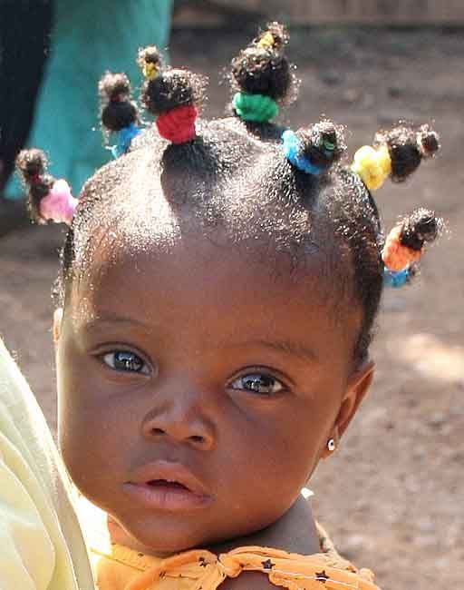 a closeup portrait of a young african american child with their hair styled into small ponytails secured with colorful hair ties. the child has brown eyes and a serious expression. an earring is visible in the right ear. the background is blurred.