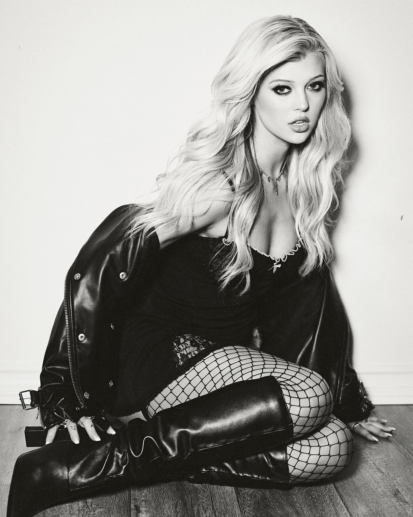 a black and white portrait of taylor momsen sitting on a wooden floor, wearing a black lingerie, fishnet stockings, black leather boots and a leather jacket. she has long blonde wavy hair and is looking directly at the camera.