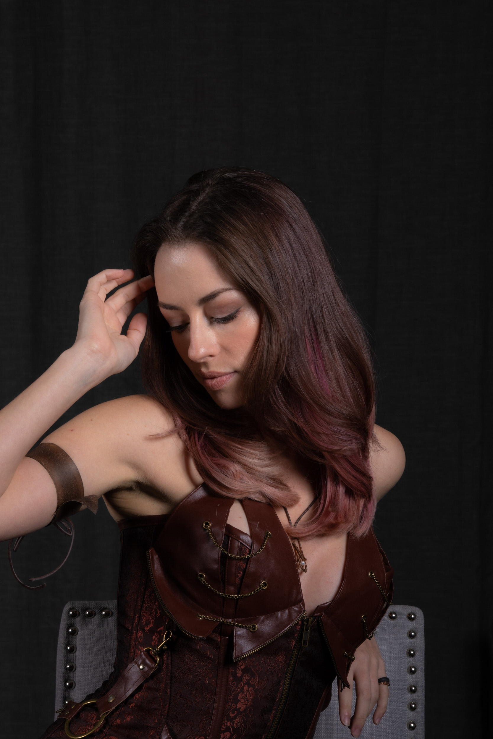 a brunette woman with pink highlights in her hair sits facing slightly away from the camera with her eyes cast down. she is wearing a brown steampunkstyle corset top with a leatherlike texture. the corset has buckles, straps, and metal accents. she is also wearing a leather cuff on her left arm. the background is a simple black backdrop, and the lighting is soft and flattering.  the overall mood of the image is mysterious and alluring.