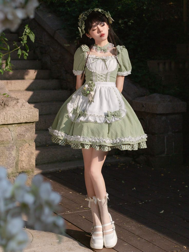 a young woman wearing a green and white lolita fashion dress is standing outdoors on a stone path in a garden. she is posing for the camera with her eyes closed and a slight smile. the dress is a square neck with puff sleeves and a full skirt. there is a white apron layer over the dress with green trim and bows. the woman has on white socks and shoes, and her hair is styled with a green headband. the background is lush and green, with stone steps and foliage visible. the lighting is natural and soft, casting gentle shadows and highlighting the details of the dress and the setting.
