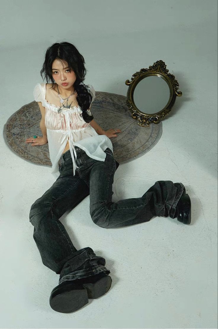 a fullbody shot of a young asian woman posing on a rug. she's wearing a sheer white blouse with lace details, darkwash bellbottom jeans, and chunky black boots.  she's seated, leaning slightly to her side, with her legs slightly spread apart. a vintage ornate mirror rests on the rug behind her. the overall style is vintageinspired, blending romantic and grunge elements. the backdrop is a minimalist light gray studio setting.